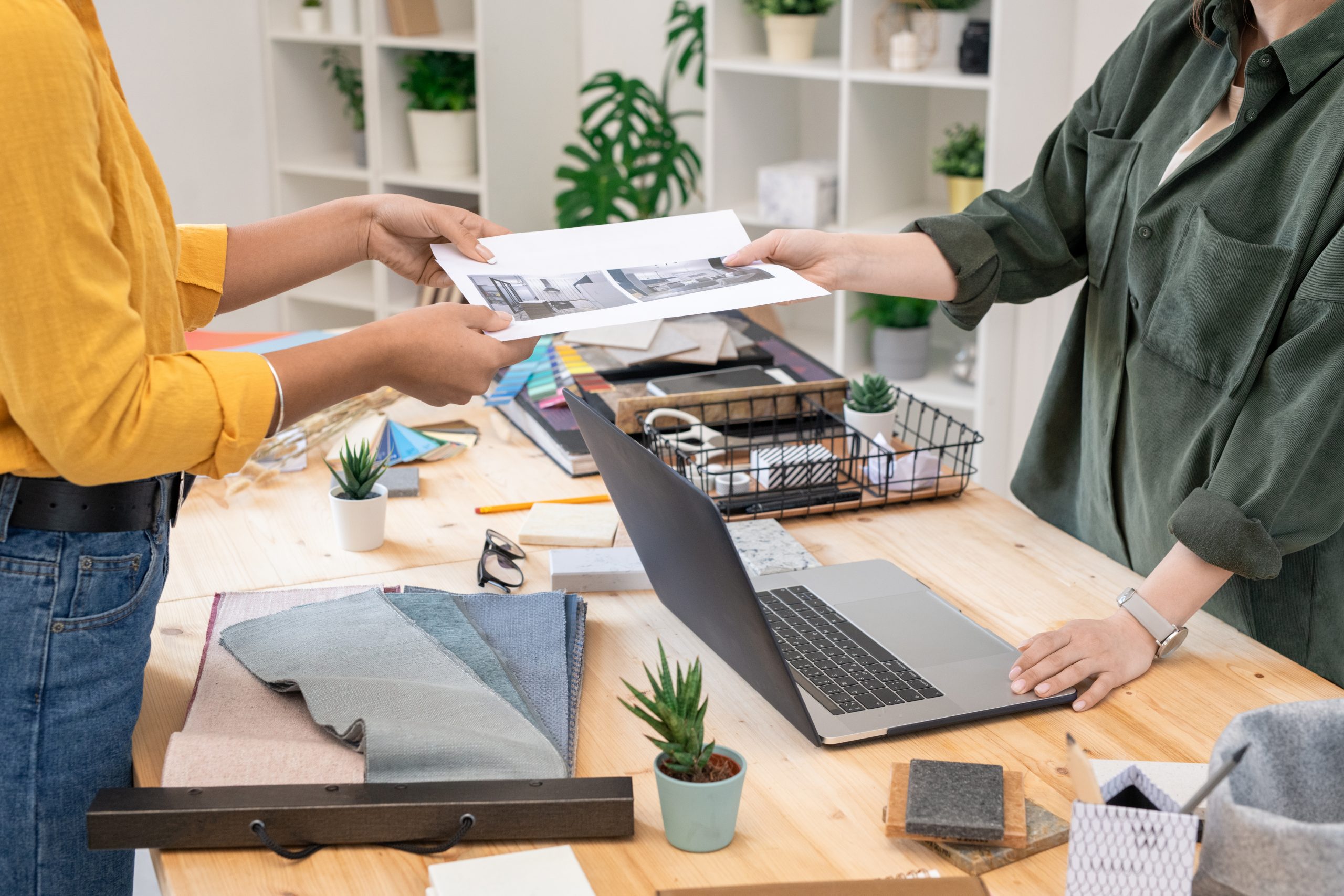 Young casual woman passing paper with photograph of room to female interior designer over her workplace while making order